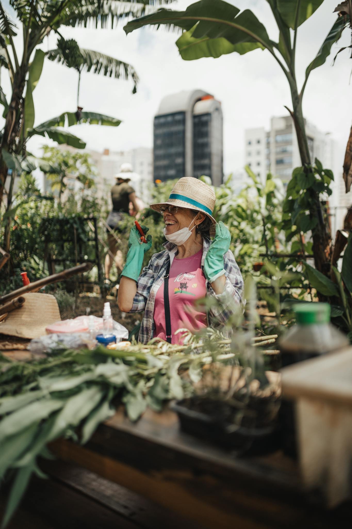 A cheerful woman gardening on a rooftop surrounded by tall green plants in an urban setting.
